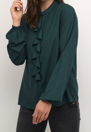 Mujer con blusa de manga larga verde oscuro con detalle de volantes al frente, combinada con pantalones negros, posando con una mano cerca de su rostro.