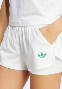 Witte sportshorts met een geplooide tailleband, zijzak en groen Adidas-logo. Gemaakt van glad, lichtgewicht materiaal met een subtiele textuur.