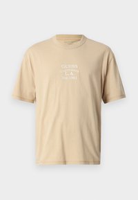 Beige bomull t-skjorte med korte ermer, med hvit brodert tekst: "GUESS CALIFORNIA L.A. Est.1981" på fronten. Rund hals.