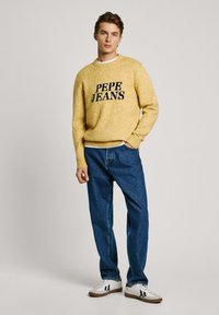 Geltonas megztinis su "PEPE JEANS" logotipu, derinamas su mėlynomis džinsinėmis kelnėmis ir baltomis sportinėmis bateliais su šviesiai rudi akcentais.