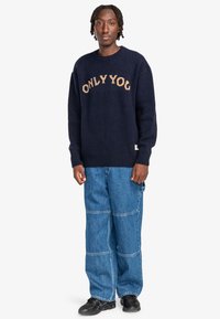 Mörkblå stickad tröja med "ONLY YOU" i beige bokstäver, i kombination med lösa blå denimbyxor och svarta skor. Strukturerad tyg, avslappnad passform.