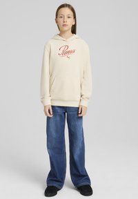 Felpa beige con logo rosso "Puma", tasca frontale e cappuccio con cordino, abbinata a jeans blu a gamba larga e scarpe nere.