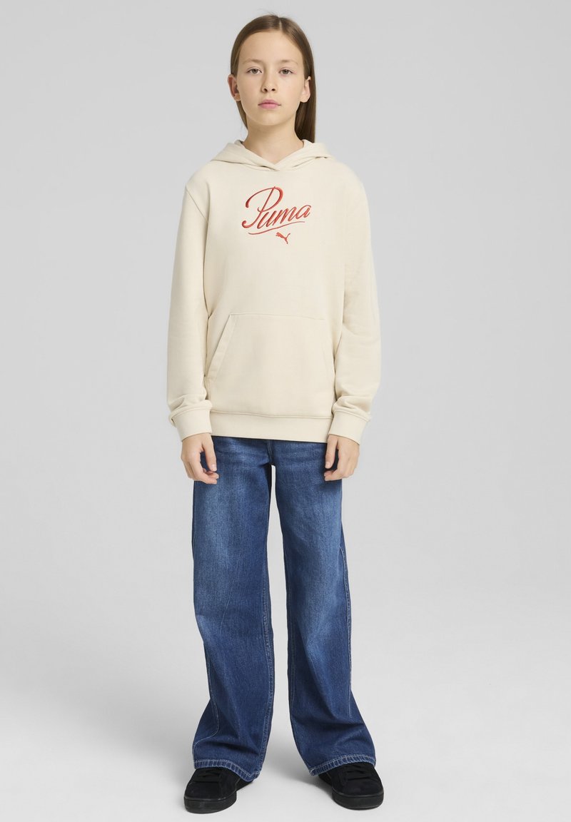Felpa beige con logo rosso "Puma", tasca frontale e cappuccio con cordino, abbinata a jeans blu a gamba larga e scarpe nere.