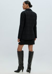 Zwarte tweed blazer met een gestructureerd patroon, getailleerd ontwerp en gestructureerde kraag; gecombineerd met een bijpassende mini rok en zwarte kniehoge laarzen.