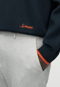 Μπλε πουλόβερ με πορτοκαλί κέντημα "Samsoe" και λεπτομέρεια μανσέτας σε πορτοκαλί χρώμα, συνδυασμένο με μαύρο-άσπρο ριγέ παντελόνι, με χέρι στην τσέπη.