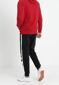 Sudadera con capucha roja, pantalones jogger negros con franjas blancas a los lados y zapatillas blancas. Tela suave con un corte relajado, costuras visibles y textura.