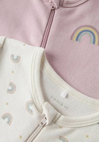 Zwei Kleidungsstücke: eines in hellrosa mit einem Frontreißverschluss und gesticktem Regenbogen, das andere in cremigem Farbton mit pastellfarbenem Regenbogenmuster und Reißverschluss.