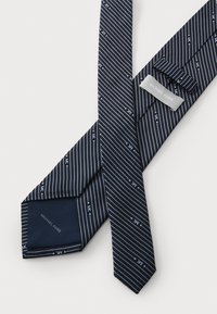 Cravate en soie bleu marine avec des rayures blanches diagonales et de petits logos 'MK'. Présente une étiquette indiquant "MICHAEL KORS" et une texture lisse.
