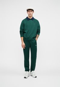Felpa verde con colletto blu navy, jogging verde abbinati e sneakers bianche. Presenta un semplice logo sul petto e una vestibilità casual.