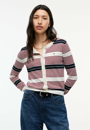 Junge Frau mit langen dunklen Haaren, die eine rot-weiß-navy gestreifte Strickjacke und blaue Jeans trägt, steht mit den Händen hinter dem Rücken.