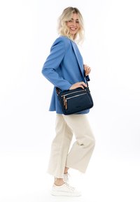 Borsa a tracolla blu navy con due tasche anteriori con zip, dettagli in pelle marrone, forma rettangolare, realizzata in tessuto resistente, indossata con abbigliamento casual.