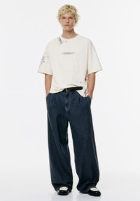 Tricou oversized crem cu un design de stropi, asortat cu blugi negri cu picioare largi și încălțări negre cu detalii albe, persoană în picioare.