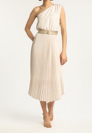 Femme portant une robe midi plissée beige à une épaule avec une ceinture dorée et des sandales dorées à lanières, debout devant un fond uni.
