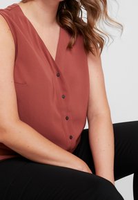 Blouse à manches courtes et col en V de couleur rouille, avec une fermeture à boutons sur le devant et une poche unique. Portée avec un pantalon noir. Texture lisse.