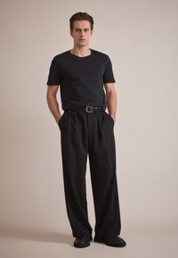 T-shirt noir à manches courtes associé à un pantalon noir taille haute. Le pantalon présente des plis et une ceinture avec une boucle en argent. Chaussures noires texturées.