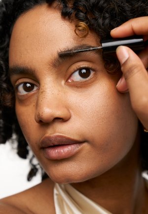Personne appliquant du gel à sourcils avec un pinceau noir, présentant une peau brun moyen, des cheveux bouclés foncés et une texture naturelle et lisse des traits du visage.