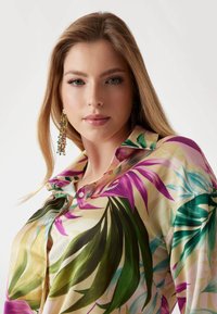 Bloemenprint shirt met levendige paarse en groene bladeren op een zachte beige achtergrond, met een klassieke kraag en knoopdetail.