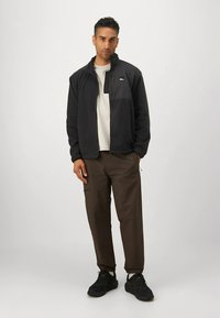 Schwarze Fleecejacke mit hohem Kragen, grauem T-Shirt darunter, braune Hose und schwarze Turnschuhe. Einfaches Design ohne auffällige Muster.