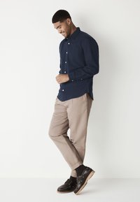 Marinblå skjorta med knappar och en bröstficka, krämfärgade byxor och svarta snörskor med texturerad sula, mot en vit bakgrund.