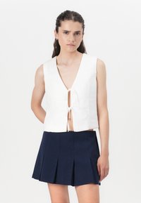Witte cropped top met een diepe V-hals en een striksluiting aan de voorkant, gecombineerd met een marineblauwe geplooide minirok. Soepele textuur, losse pasvorm.