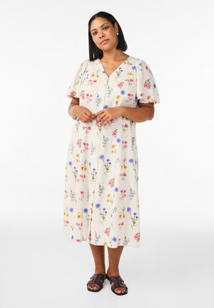 Vrouw staand, gekleed in een crèmekleurige midi-jurk met roze, blauwe en gele bloemenprint en zwarte open-toe sandalen met studs.
