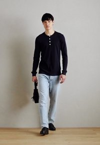 Svart langermet henley-skjorte med tre hvite knapper, lyseblå jeans og svarte sko. Personen holder en liten svart veske.