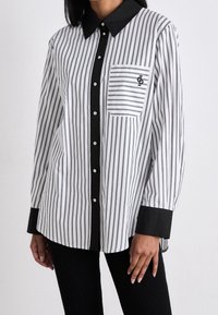 Chemise à rayures noire et blanche avec col classique, détail de poche, boutons en argent et poignets noirs contrastants.