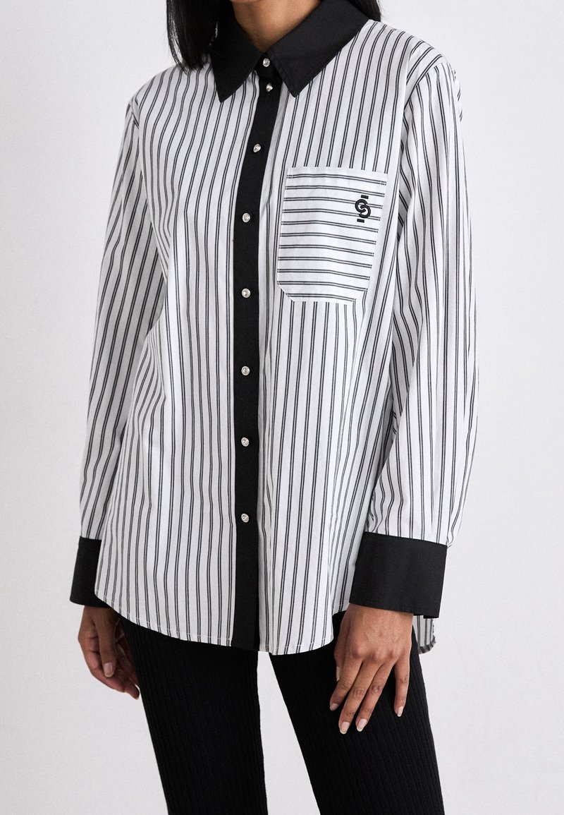Chemise à rayures noire et blanche avec col classique, détail de poche, boutons en argent et poignets noirs contrastants.