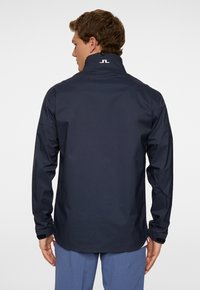 Man som bär en marinblå vattentät jacka med hög krage och logga på ryggen, ihop med blå byxor, stående mot en neutral bakgrund.