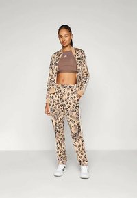 Nő áll, leopárdmintás tréningdzsekiben és nadrágban, barna crop toppal és fehér sportcipővel, egy sima világos háttér előtt.