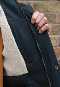 Veste noire avec une doublure en polaire beige douce, dotée d'une fermeture à glissière et d'une poche extérieure visible sur le côté gauche.