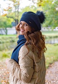 Căciulă tricotată de culoare albastră închisă, cu un pom-pom din blană sintetică și un accent decorativ din metal; asociată cu o eșarfă albastră închisă și o geacă pufoasă bej.