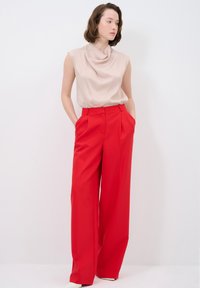 Beige drape-top zonder mouwen, gecombineerd met hooggewaist, wijde rode broeken met zijzakken en scherpe plooien.