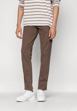 Personne portant un pantalon droit marron, une chemise à manches longues rayée beige et blanche et des baskets blanches, debout devant un fond uni.
