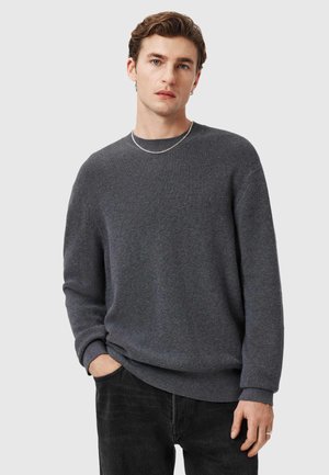 Szary, teksturowany sweter dzianinowy z okrągłym dekoltem i długimi rękawami, zestawiony z ciemnymi dżinsami. Subtelne logo na piersi.