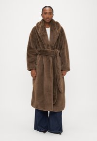 Hnedý župan z umelej kožušiny so širokým golierom, opaskom v páse a dlhými rukávmi, doplnený rozšírenými denimovými nohavicami. Hladká textúra po celej ploche.