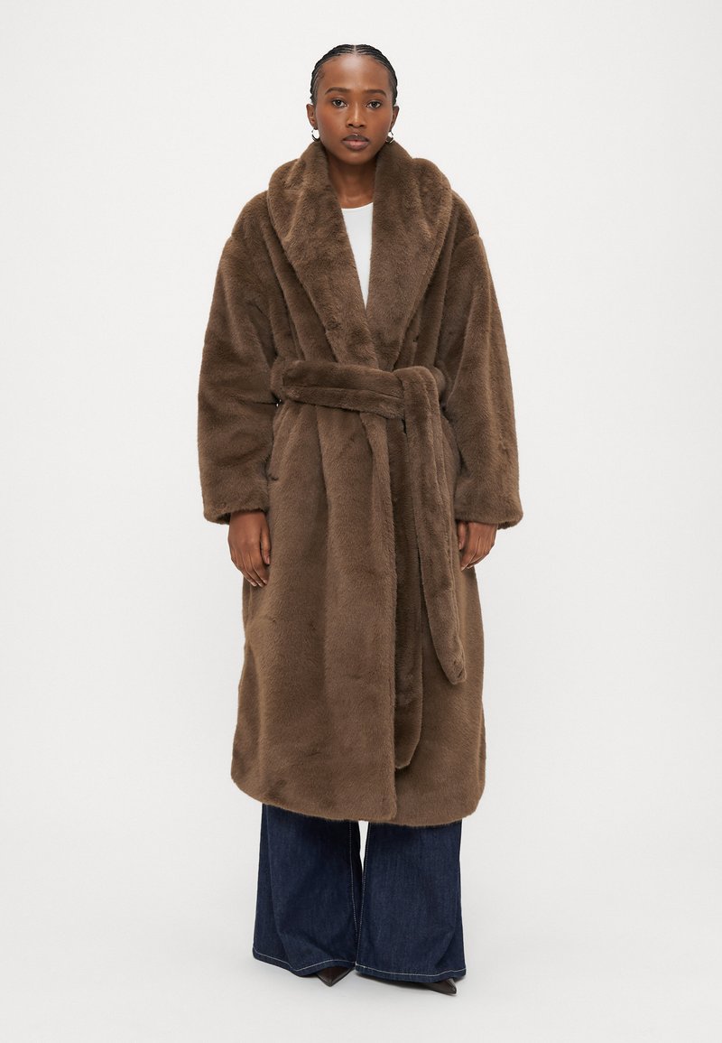 Hnedý župan z umelej kožušiny so širokým golierom, opaskom v páse a dlhými rukávmi, doplnený rozšírenými denimovými nohavicami. Hladká textúra po celej ploche.