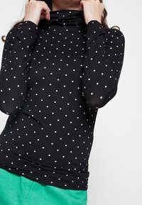 Svart långärmad polokrage med vita prickar, med en figurnära design och mjukt tyg. Stylen är matchad med gröna kordbyxor.