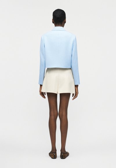 Άτομο που στέκεται με την πλάτη γυρισμένη, φοράει ανοιχτό μπλε κοντή ζακέτα, μπεζ σορτς ψηλόμεσα και φλατ παπούτσια με σχέδιο λεοπάρ, σε λευκό φόντο.