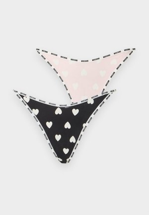 Femme debout pieds nus portant un soutien-gorge noir et un bas de bikini noir avec des motifs de cœurs blancs sur un fond clair uni.