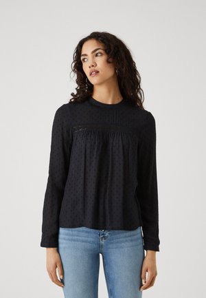 Black long-sleeve blouse with textured dot pattern and lace detail across the chest; relaxed fit with round neckline. Paired with blue jeans.