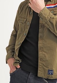 Camicia leggera color verde oliva, con bottoni e due tasche sul petto, con strisce sulle spalle rosse, bianche e blu in contrasto, indossata sopra un maglione nero.