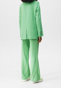 Groene linnen suit met een getailleerde blazer met een rugventilatie en bijpassende wijde broekspijpen. Eenvoudige, gestructureerde stof met contrasterende knopen.