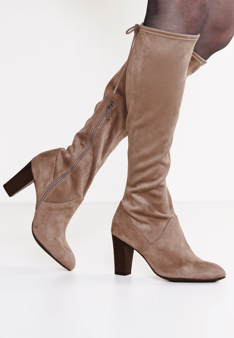 Botas de ante beige hasta la rodilla con puntera redondeada y tacón apilado. Presentan un cierre de cremallera lateral y un lazo decorativo en la parte superior del caña.