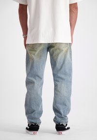 Persoon in een wit t-shirt, lichtblauwe vervaagde jeans, zwart-witte sneakers, staand met hun rug naar de camera tegen een effen achtergrond.