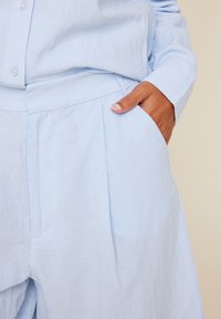 Lyseblå linbluse med matchende høyt liv bukser. Plisserte detaljer og sidelommer kan sees, noe som fremhever en avslappet passform og tekstur.