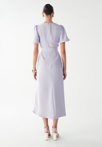 Lila midi obleka s puhastimi rokavi, prilegajočim pasom in zadrgo na hrbtu. Oblečena s belimi sandali na visoki peti, gladko blago in rahlo sijaj.