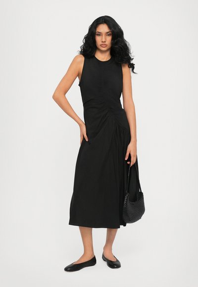 Black sleeveless dress featuring gathered detailing at the waist, ankle length, paired with black ballet flats and a woven handbag.