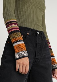 Närbild av en person som bär en grön ribbad tröja med flerfärgade mönstrade muddar och svarta högmidjade jeans.