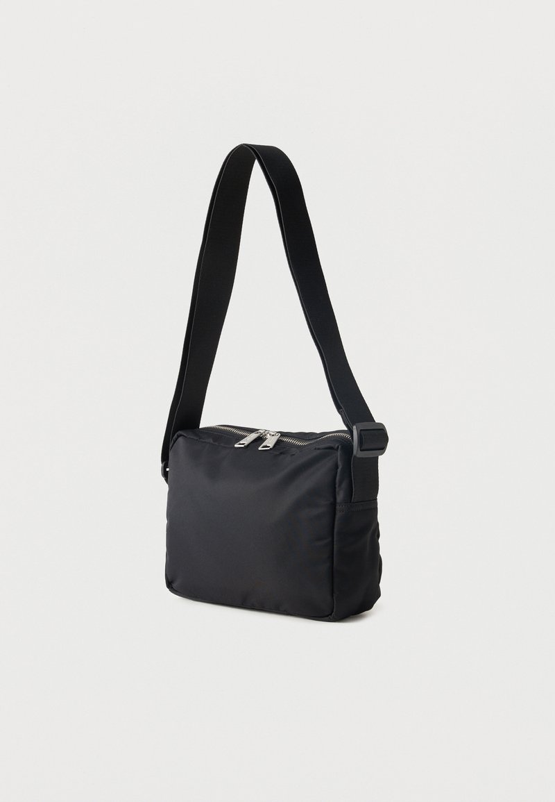 Sac bandoulière rectangulaire noir avec large sangle ajustable et double fermeture éclair argentée sur le dessus.